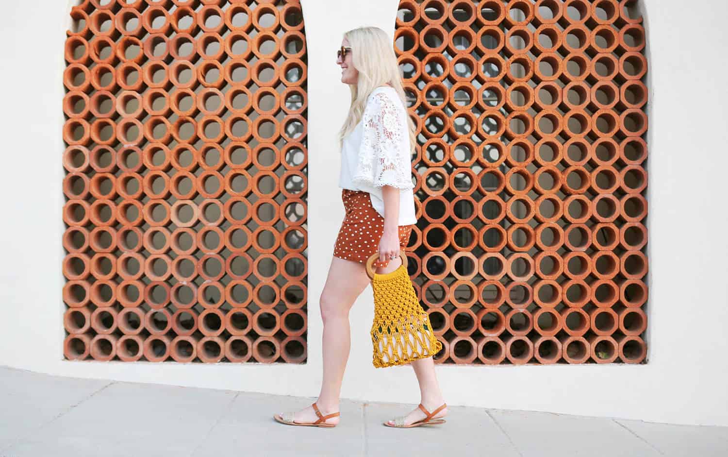 a woman wearing a white long sleeve shirt, red with white polka dots shorts, and sandals holding a yellow macrame handbag